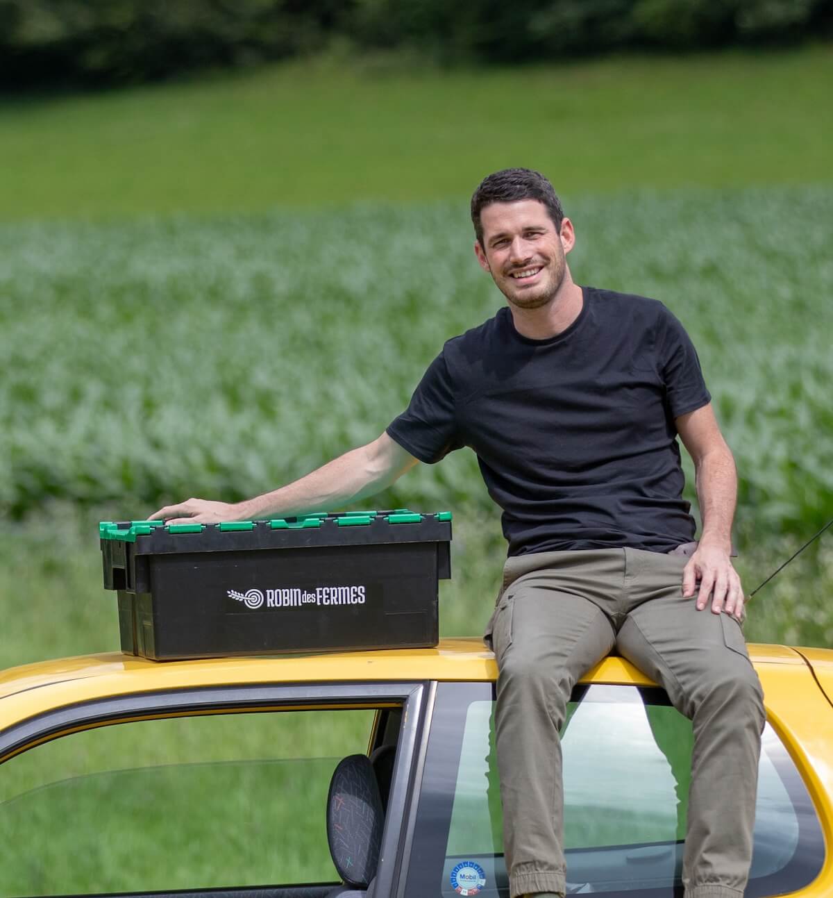 Tanguy, assis sur une voiture jaune avec une caisse noire de Robin des Fermes sur le toit, souriant devant un champ verdoyant.