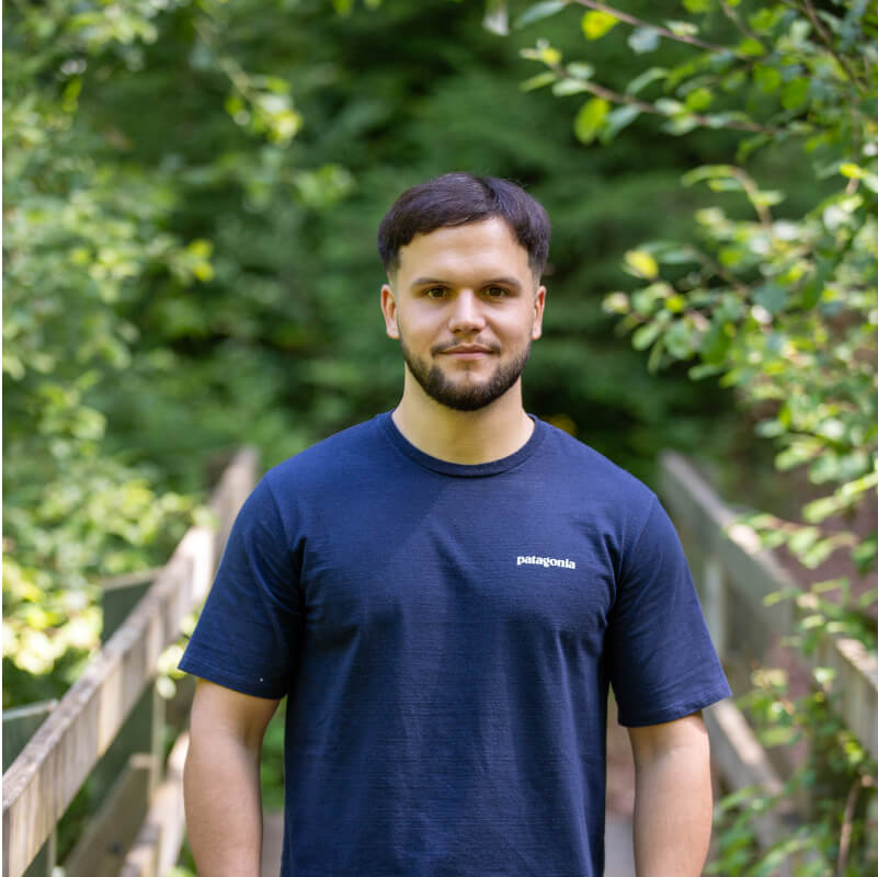 Portrait d'Alex portant un t-shirt bleu et se tenant sur un pont en bois entouré de verdure