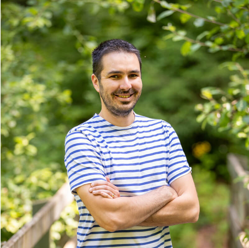 Portrait de Fabien portant un t-shirt rayé blanc et bleu, bras croisés, avec un fond de verdure.