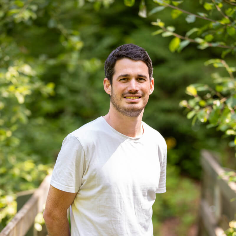 Portrait de Tanguy portant un t-shirt blanc, posant en extérieur avec un fond de verdure.